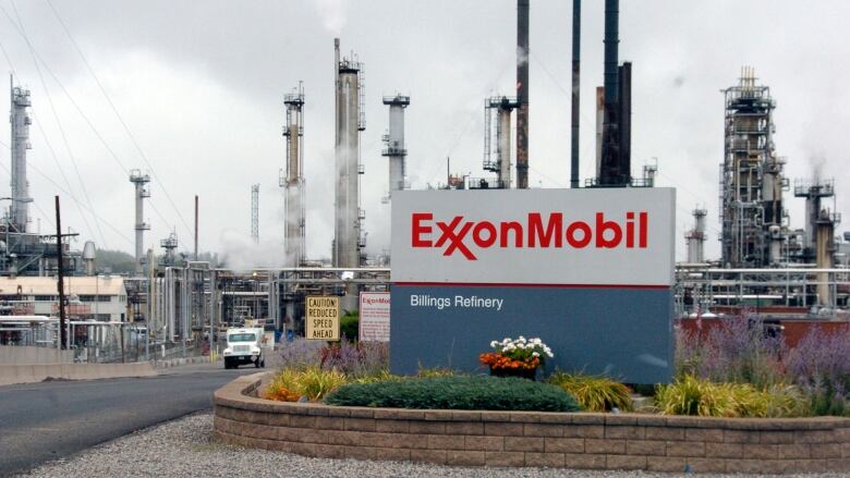 A sign for Exxon Mobil sits in the foreground. Behind it, smoke stacks rise into the sky and a large truck drives out of the refinery.