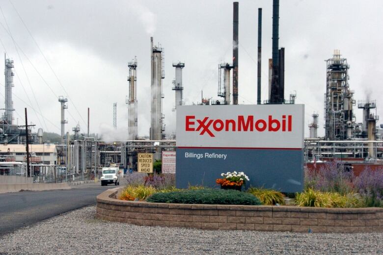 A sign for Exxon Mobil sits in the foreground. Behind it, smoke stacks rise into the sky and a large truck drives out of the refinery.