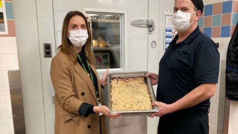 A woman in tan on the left and a man wearing black on the right hold up a pan of blueberry crisp to face the camera. 