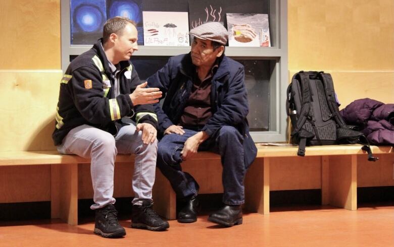 A pair of men sit, facing each other, on a bench. The man on the left has lifted his right hand as he speaks, while the man on the right is looking at the camera out of the corner of his eye.