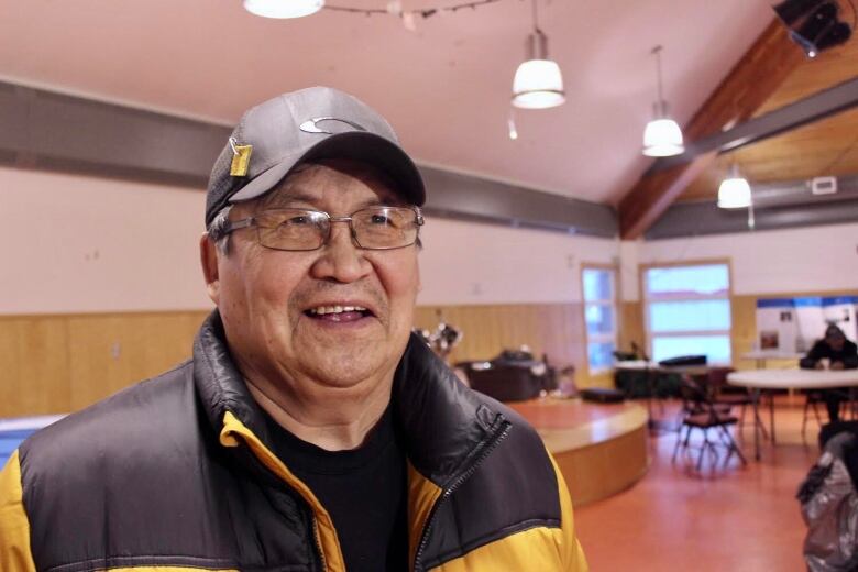 Headshot of a man wearing glasses, a black baseball cap with a piece of leather pinned to it, and a black and yellow coat.