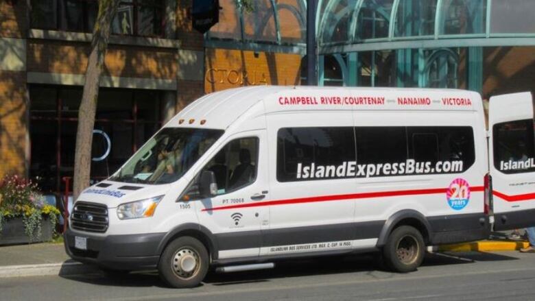 A white passenger van parked outside a brown building with white lettering that says Islandexpressbus.com along the window and Campbell River/Courtnenay-Nanaimo-Victoria in red letters above the windows.