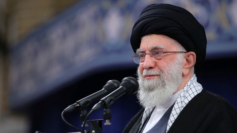 The ayatollah is in front of two microphones. The background is out of focus and largely blue.