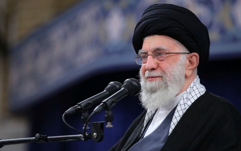 The ayatollah is in front of two microphones. The background is out of focus and largely blue.