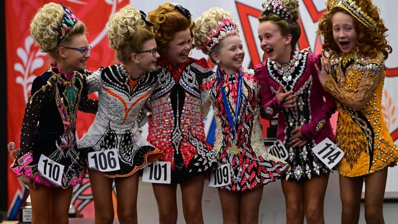 A group of young girls in elaborate costumes cheer. They are wearing numbers and all have wigs on.