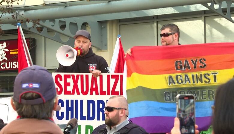 Two men hold signs in protest of a Drag Queen story hour event. They are flanked by police officers and people holding phones.