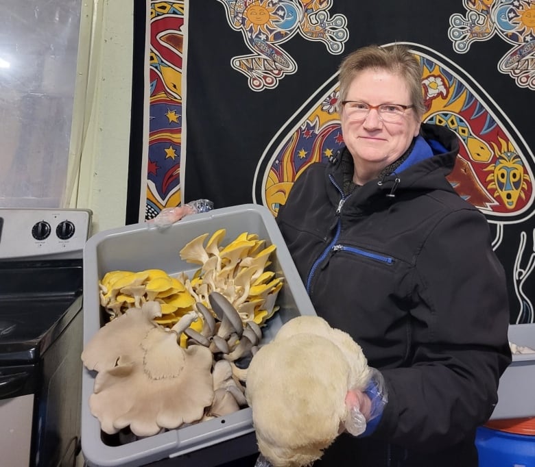 A middle-aged woman with short hair and glasses holds a container full of large edible mushrooms. Some are yellow and some are white. 