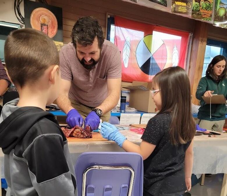 A man cuts into a muskrat in front of two young students. 