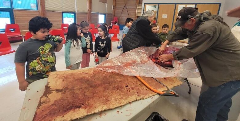 A man cuts up a piece of moose meat while children look on.