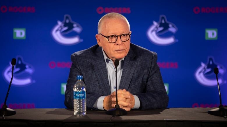 A man in a dark suit and white open-collared shirt sits at a desk in front of a microphone with a plastic water bottle beside him, waiting for questions. Behind him is the Vancouver Canucks large-C-and-orca logo on a blue background.