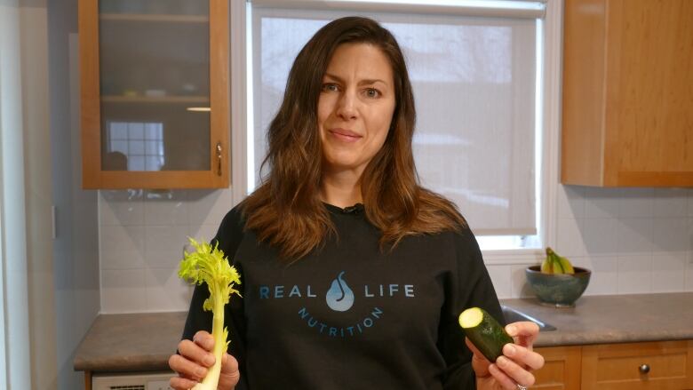 A woman standing in her kitchen holding up the last bits of a stalk of celery and a cut piece of zucchini. 