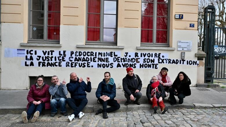 Eight people sit on the sidewalk in front of a building, where a message has been written in big block letters on the wall. The message in French reads: 'Here lives pedocriminalJ. Rivoire. The Canadian justice is asking for his extradition. Frances is refusing. We are ashamed.'