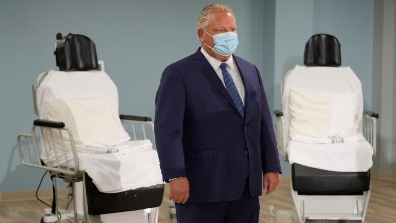 Ontario Premier Doug Ford standing, with two medical chairs behind him. 