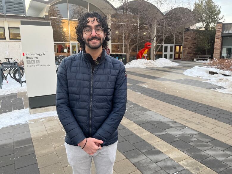 Rezaei-Afsah stands in front of the Kinesiology building in a dark blue, light winter jacket and grey sweatpants. His curly black hair falls just above his black-rimmed glasses. 