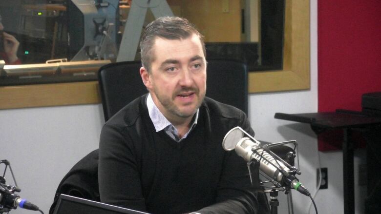 A man wearing a black sweater sits in a radio studio in front of a microphone.