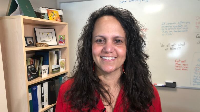 A woman with curly hair wearing a red shirt smiles in front of a whiteboard.