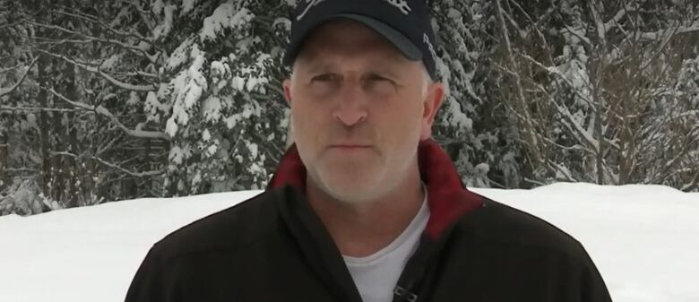 A man in a black jacket and cap stands outside with snow and trees behind him.