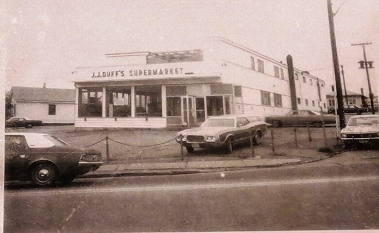 A black and white photo of a grocery store with cars in its parking lot. The sign reads J.J. Duffs Supermarket.