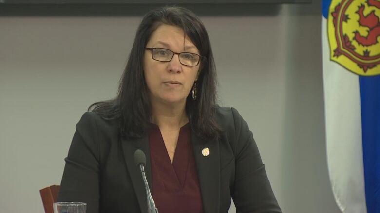 A woman sits at a desk with a microphone and a Nova Scotia flag behind her.