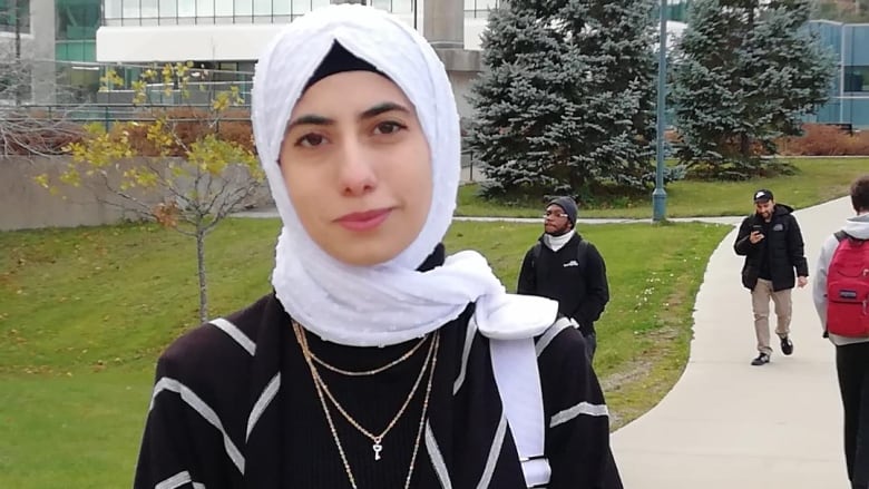 A woman wearing a white headscarf stands outside on a paved walkway near green grass. 