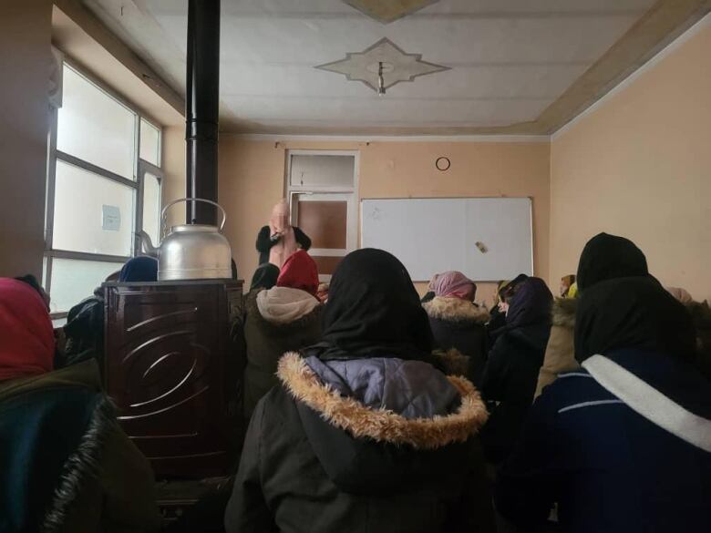 A teacher stands in front of a classroom of girls wearing winter jackets. Her face is blurred.