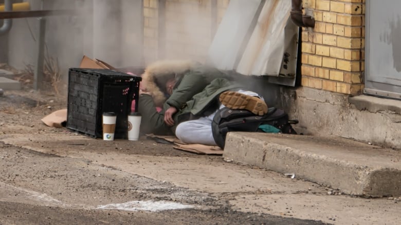 A person wearing a parka, with a hood pulled up around their head, lies on the concrete in an alleyway.