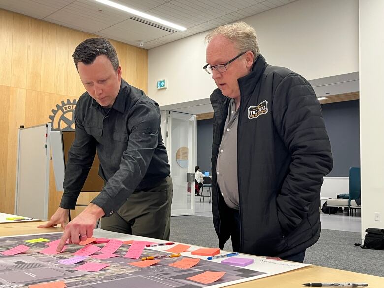 A man points to a sticky note on a huge map while a second man looks on. 