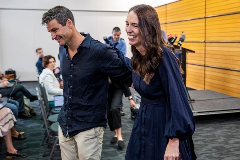 A man and woman, both wearing navy blue and smiling, walk arm in arm away from a podium.