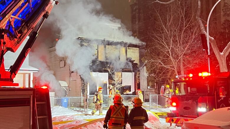 Smoke pours out of a two-storey house in the centre of the photo. Firefighters stand in the foreground wearing helmets and protective coats. Much of the photograph is highlighted in red from the emergency lights on fire trucks.