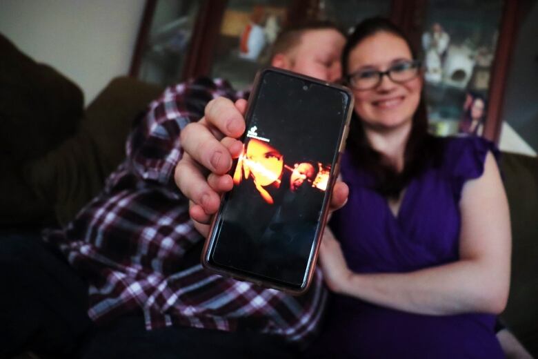 A man kisses a woman on her cheek as they sit on a couch and hold up a cell phone displaying an image of them performing.