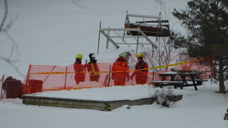 Four people wearing hard hats and high visibility construction suits stand on the ice next to a dock, surrounded by orange construction fencing and oil drums 