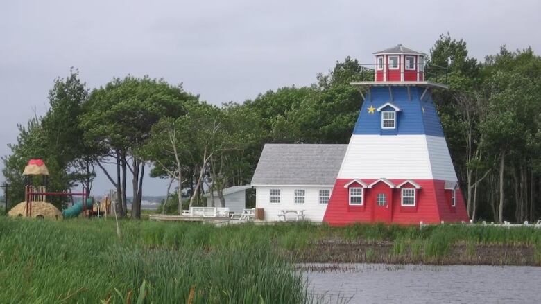 A lighthouse in the colours of the Acadian flag, with a pond in front of it, a white building behind it and a playground to its right.