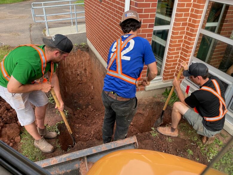 Three men carrying shovels dig around the foundation of a house. 