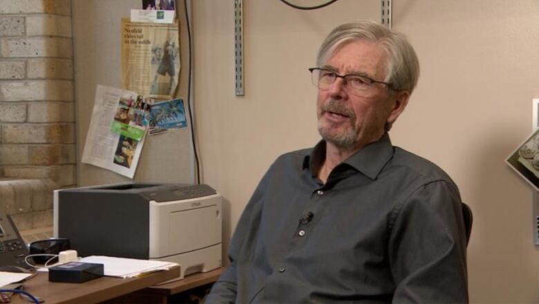 A man with grey hair clad in a grey shirt is seen speaking. A printer sits next to him.