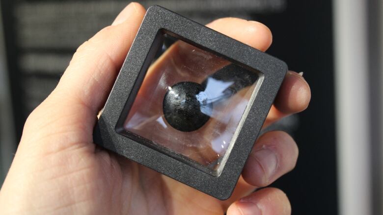 A small black flat sphere inside a little display box held in a hand.