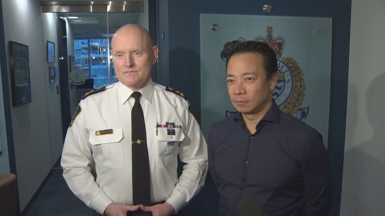 Two men, one wearing a police uniform, look at reporters asking them questions.