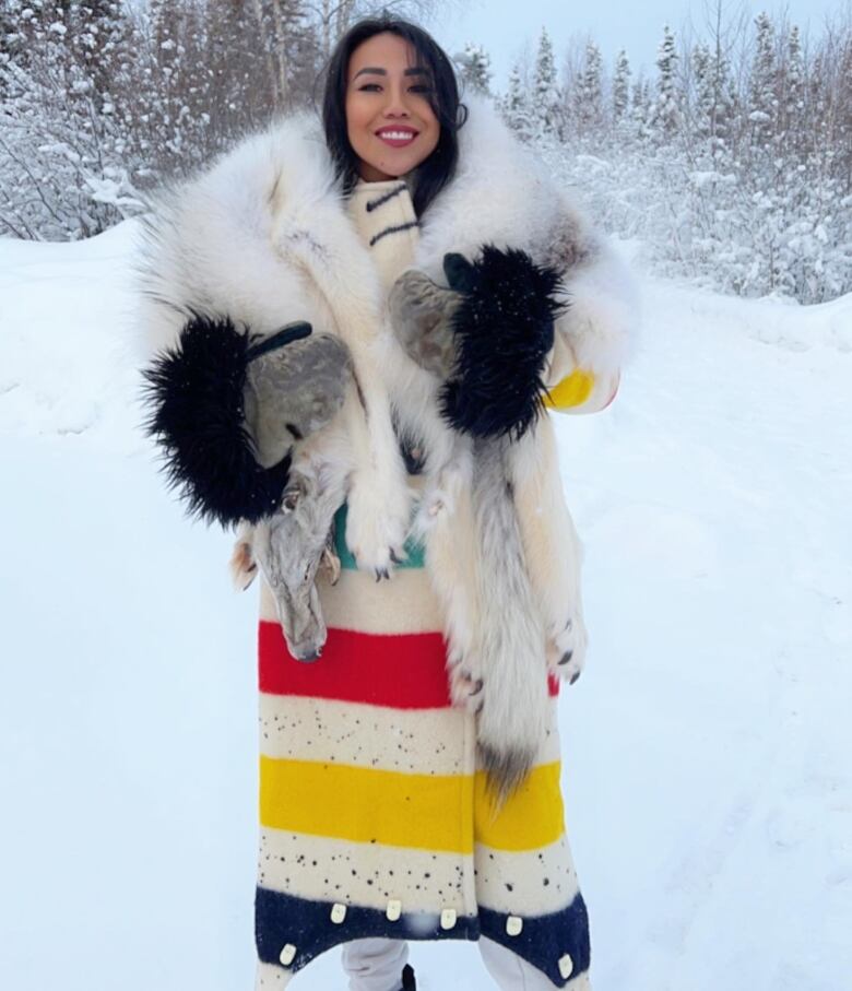 A woman standing in a snowy landscape wearing an Indigenous style coat. It's white with fur and has yellow, green and red stripes on it.