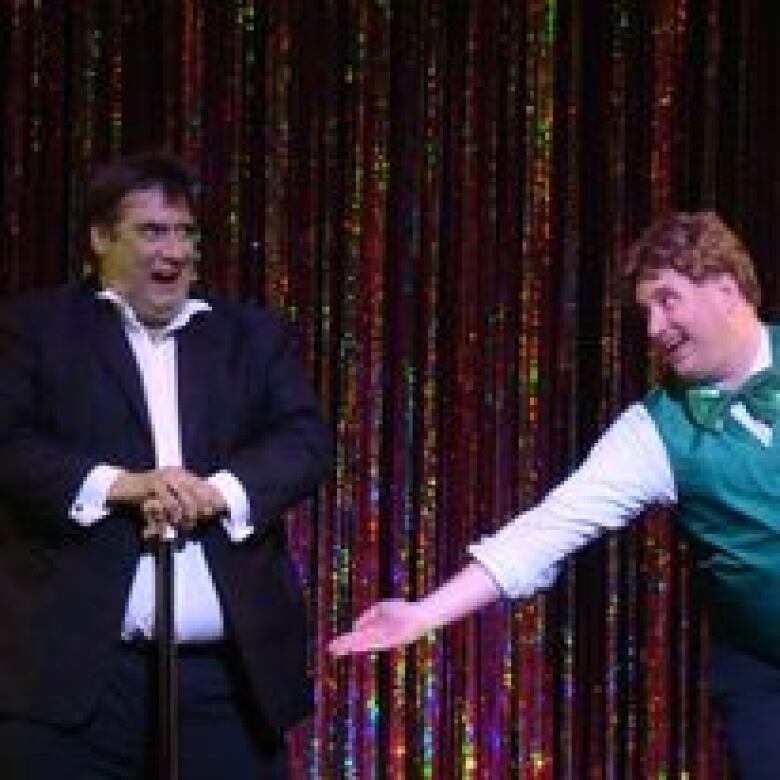 Two men on stage - the one on the left has dark hair and is wearing a black suit with a white dress shirt - and the man on the right has reddish hair, a white long sleeve shirt and green vest with a green bowtie.