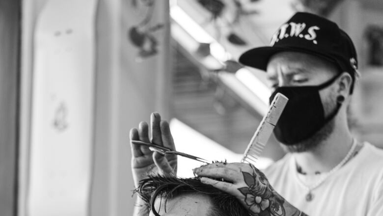 A person wearing a mask and baseball cap cuts another person's hair.