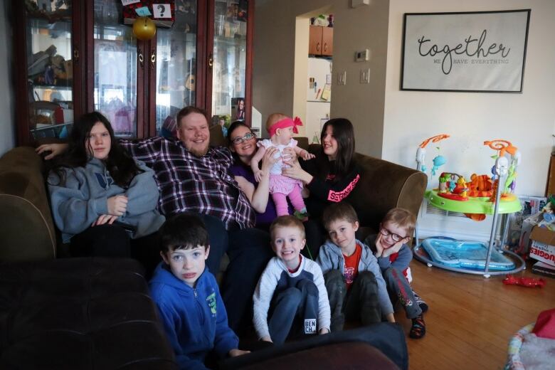 A large family sits on and in front of a brown couch in a suburban home.