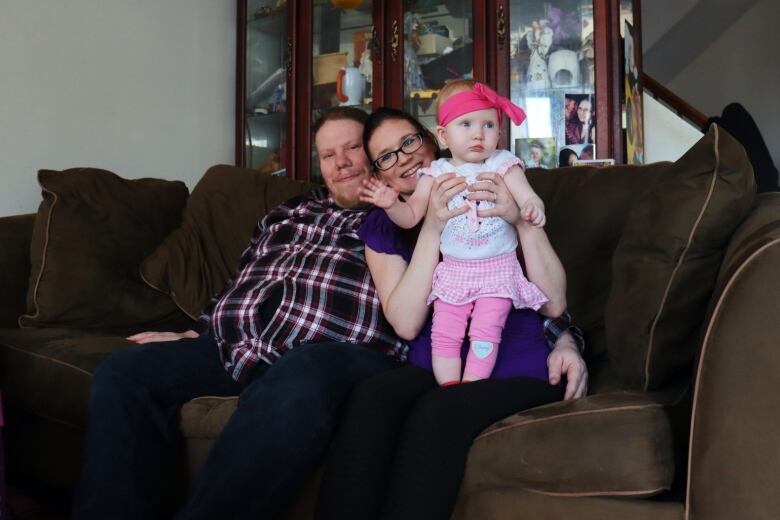 A man and woman sit together with their baby daughter on a couch, with the woman holding the baby up and having her wave to the camera.