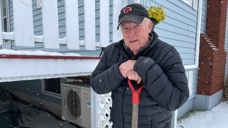 An elderly man leans on a snow shovel in his yard, standing next to an electric heat pump on his home.