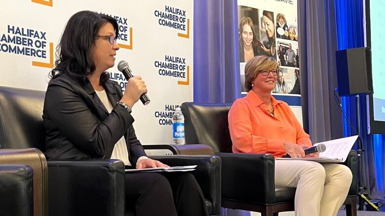 Two women sit on chairs on a stage. One holds a microphone.