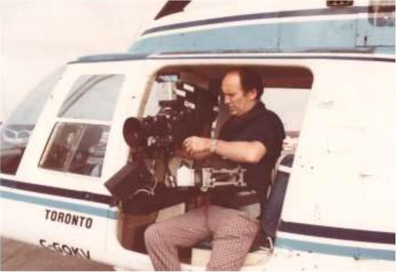 A man holds a camera rig while seated on the perch of an aircraft.