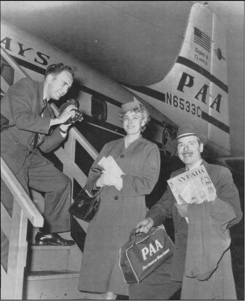 A black-and-white picture of a man with a camera boarding a plane labelled 'PAA'. A woman is in the centre, with a man carrying a newspaper to the right.