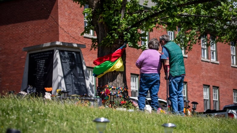 Two people look at a memorial of flags and ornaments draped on a tree, standing outside a large brick building.