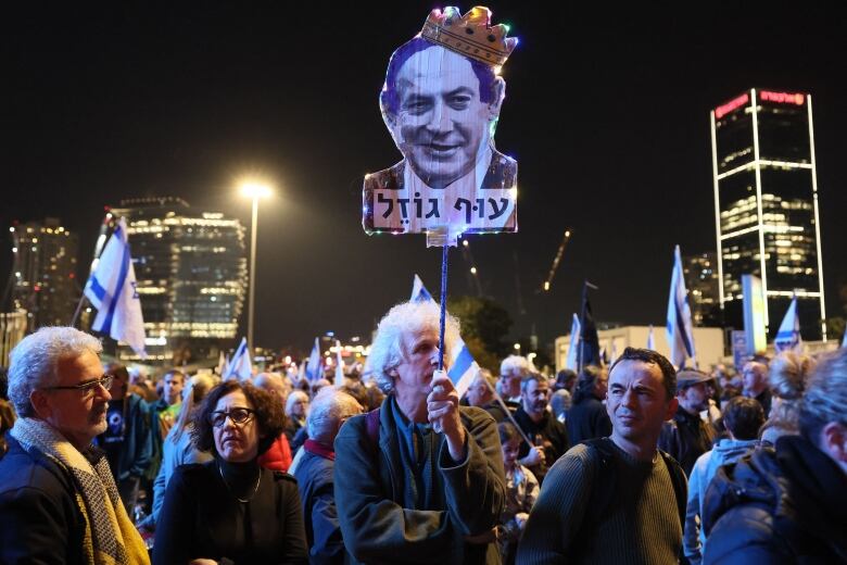 A man with long, white, curly hair holds a sign with the face of a smiling man wearing a crown.