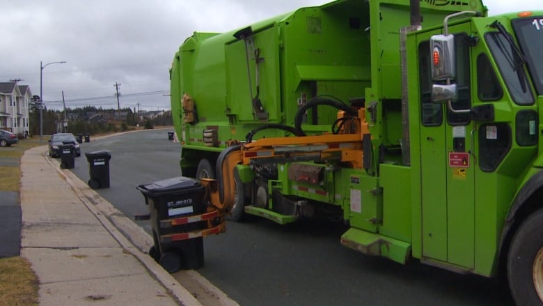 A garbage truck, on the right side of the frame, is parked on the street. A claw extended from the truck is about to close around a garbage container positioned against the curb. In the background, more garbage cans can be seen lined up against the curb.