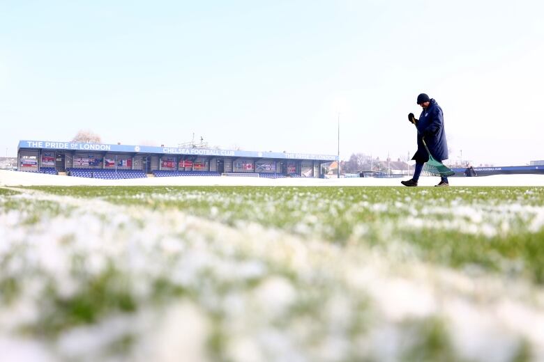 The picture shows a ground-level look at a small soccer stadium with ice in the field and a man walking with a tool to help him remove some of the ice. A small stand is seen at the far back and it reads 