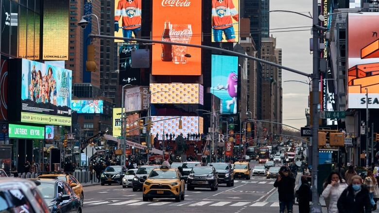 Cars, led by a yellow taxi, move through an intersection with brightly lit billboards behind them. To the right, pedestrians wait to cross a crosswalk.